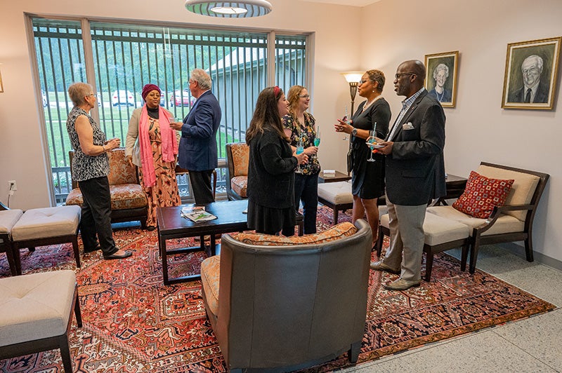 Seven people standing and talking in a room with comfortable chairs and coffee tables.